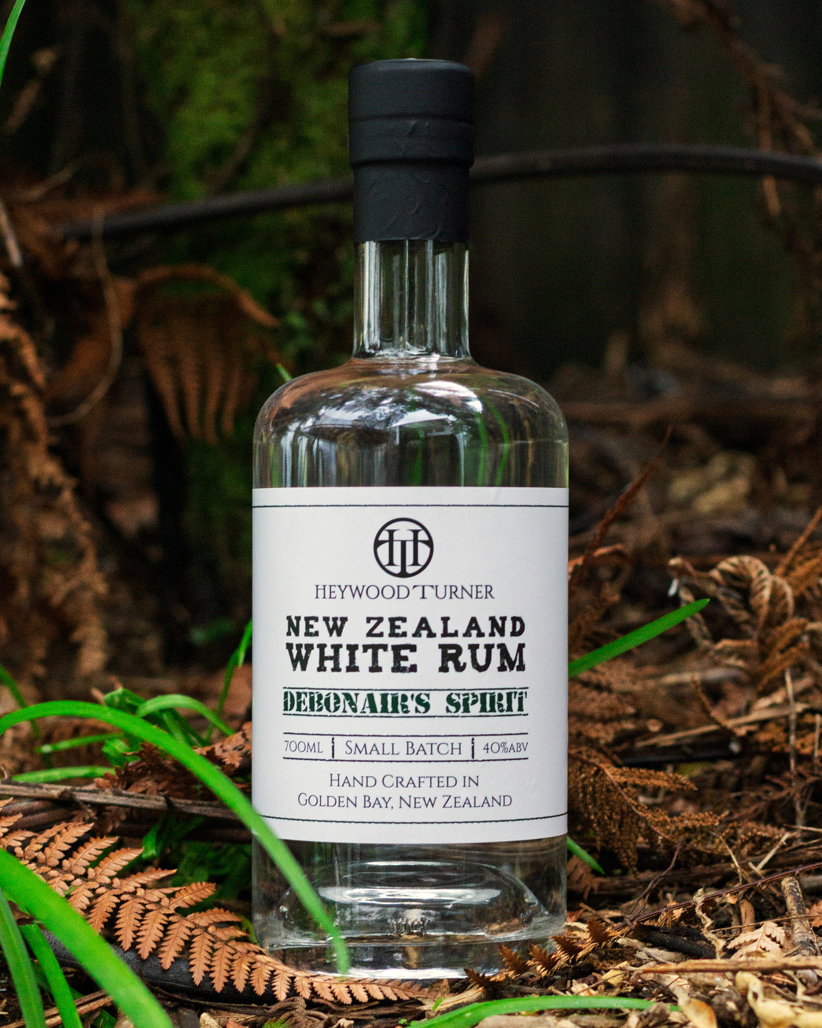 A premium white rum bottle nestled among dried brown ferns and native grass blades on a New Zealand forest floor.