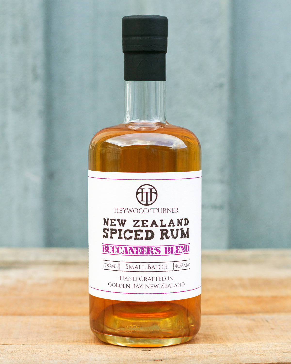 A bottle of Heywood Turner New Zealand Spiced Rum on a wooden surface with a gray textured background. The bottle contains amber-colored rum and has a white label with black and purple text.